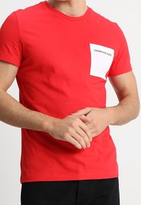 Czerwona bawełniana koszulka z okrągłym dekoltem i krótkimi rękawami. Posiada białą kieszeń z napisem "CALVIN KLEIN JEANS".