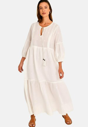 Femme portant une robe longue blanche à manches longues avec des détails brodés et un lien avec des pompons, assortie à des sandales ouvertes marron, debout devant un fond uni.