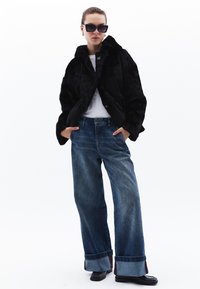 Zwarte nep bontjas met een kraag, gedragen over een wit shirt. Lichtblauwe wijde jeans met omgeslagen pijpen. Zwarte schoenen. Donkere zonnebril.