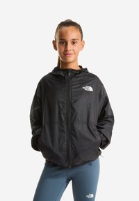 Schwarze Zip-up-Jacke mit Kapuze, aus leichtem Material. Verfügt über ein Logo auf der Brust und elastische Bündchen. Wird über blauen Sportleggings getragen.