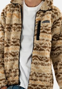 Giacca in pile marrone e crema con pattern testurizzato; presenta una cerniera nera e una tasca. Indossata sopra una t-shirt bianca.