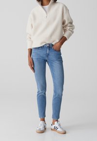 Sudadera de forro polar blanca con cremallera de cuarto, jeans de color azul claro y zapatillas blancas con rayas marinas. Textura suave con ajuste relajado.