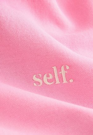 Rožinė medžiaga su minkšta tekstūra turi išsiuvinėtą žodį "self." šviesesniame atspalvyje. Dizainas yra minimalistiškas ir lygus.