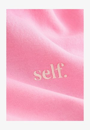 Ružová tkanina s jemnou textúrou má vyšívané slovo "self." v svetlejšom odtieni. Dizajn je minimalistický a hladký.