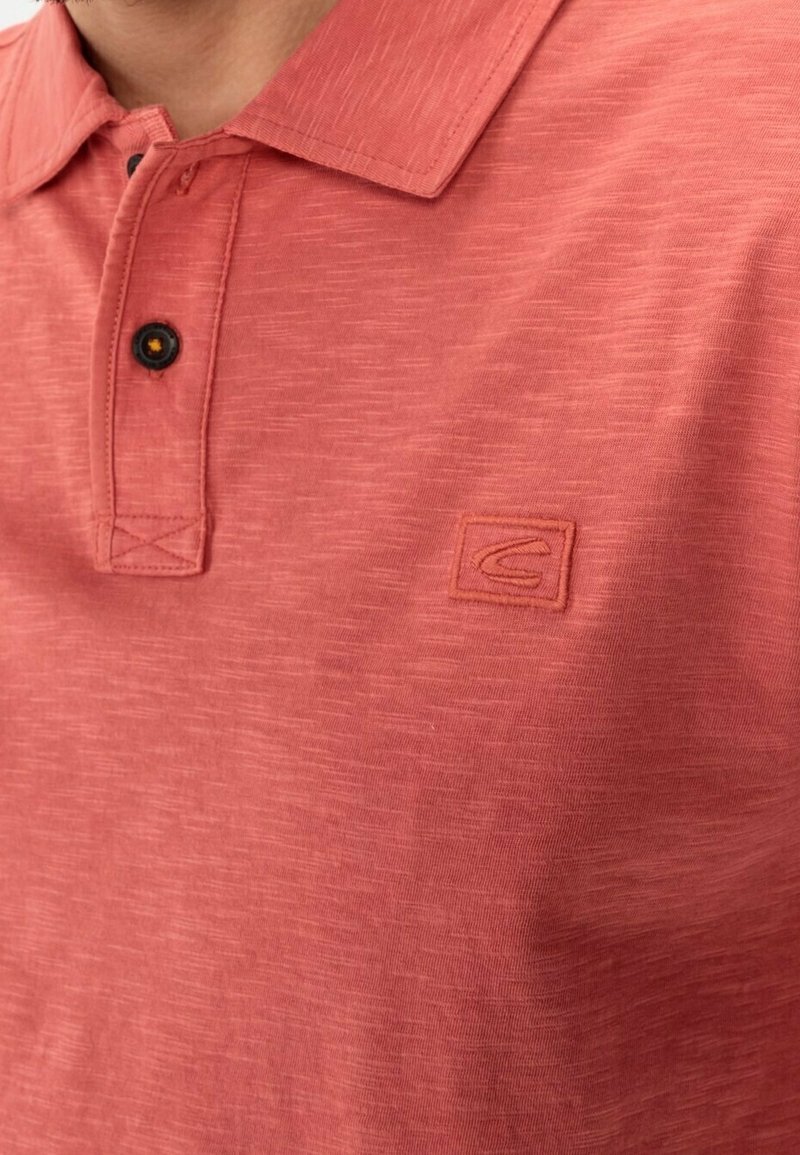 Gros plan sur une personne portant un polo corail texturé avec un col, deux boutons noirs et un petit logo brodé sur la poitrine.