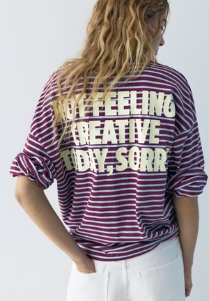 Persona con capelli lunghi che indossa una maglietta a righe bordeaux e bianche con la scritta "NON MI SENTO CREATIVO OGGI, SCUSA" sul retro.