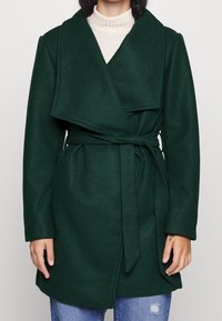 Grön ullblandad kappa med omslagdesign, breda slag och knytbälte i midjan. Mjuk textur, långa ärmar och avslappnad passform.