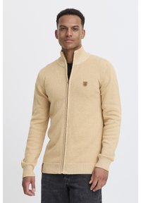 Suéter beige con cremallera, fabricado en tejido de punto texturizado, que presenta un cuello alto y un pequeño parche con logotipo en el pecho. Unos vaqueros oscuros complementan el conjunto.