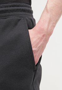 Handen instoppad i sidofickan på svarta träningbyxor med elastisk midja, synliga underarmar och handledsådror.