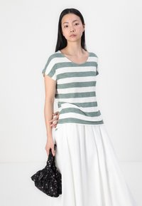 Stribet grøn og hvid T-shirt med korte ærmer og scoop-neck, parret med en hvid nederdel. Modellen holder en blomsterpræget sort taske.