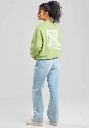 Lichtgroene oversized sweatshirt met een witte grafische print op de achterkant, gecombineerd met lichtblauwe wide-leg jeans en witte sneakers.
