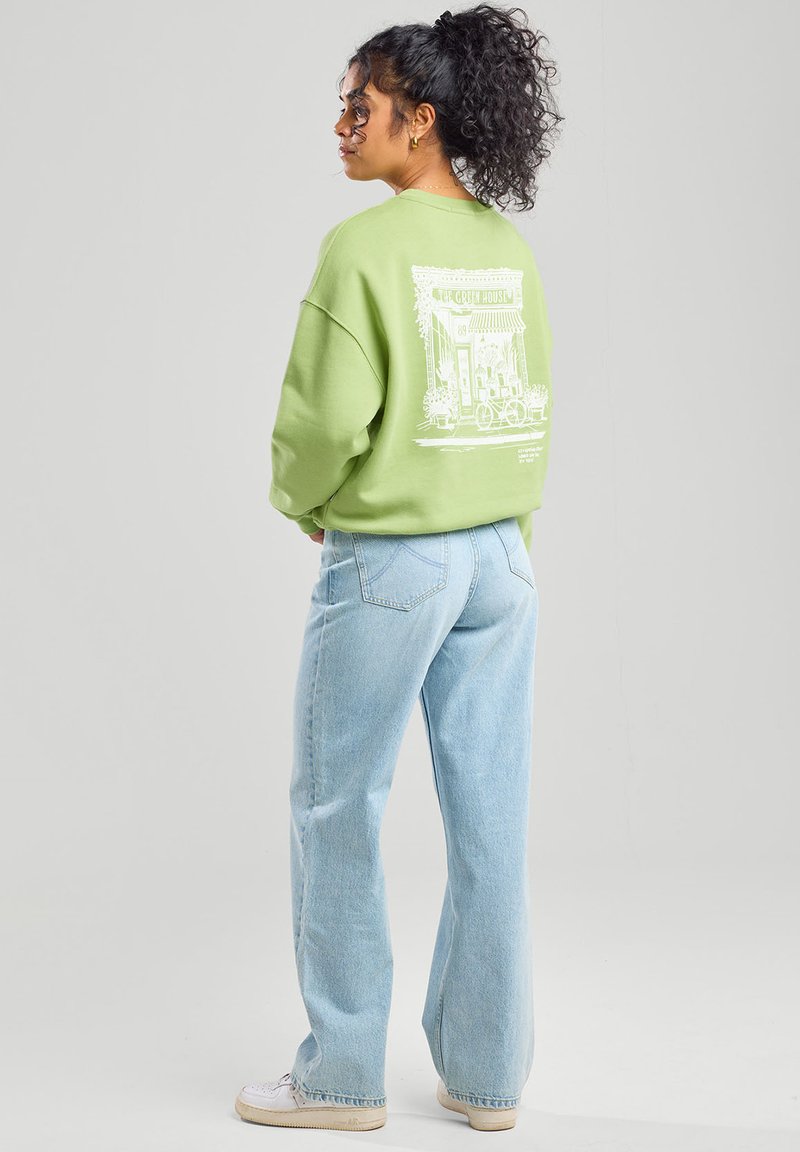Light green oversized sweatshirt with a white graphic design on the back, paired with light blue wide-leg jeans and white sneakers.