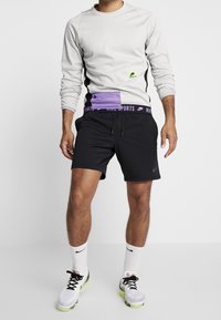 Sweatshirt gris clair avec panneaux latéraux noirs et logo vert, associé à un short noir avec une taille élastique violette. Chaussettes blanches et baskets multicolores.