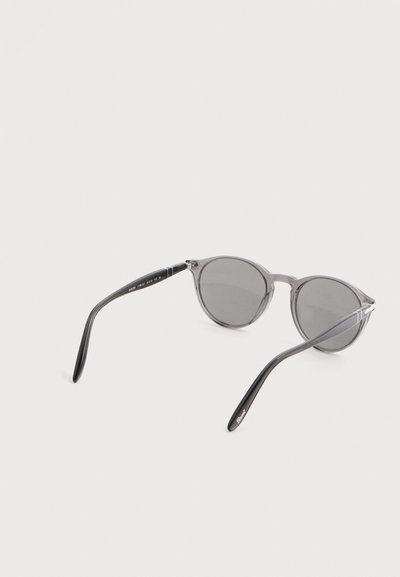 Στρογγυλά γυαλιά ηλίου με γκρι πλαίσιο και σκούρους φακούς, τοποθετημένα πάνω σε λευκή επιφάνεια, εμφανιζόμενα από πίσω.