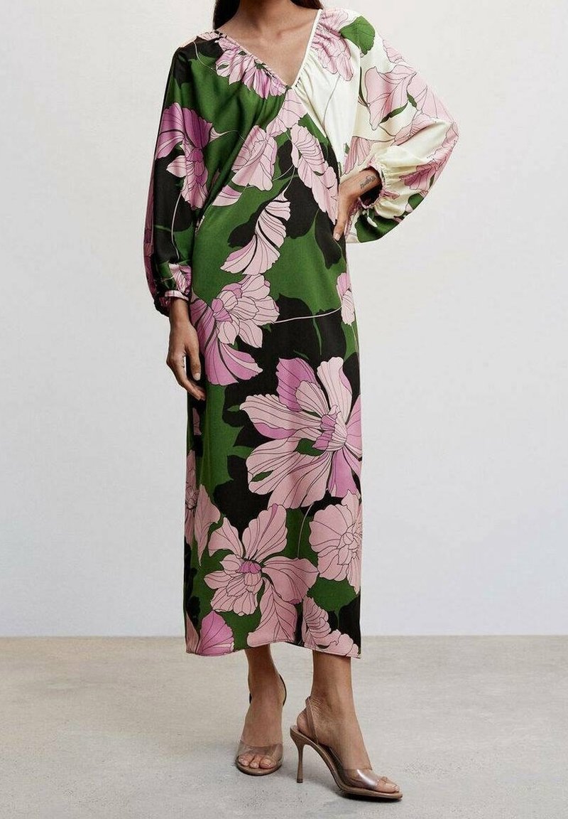 Femme portant une longue robe fleurie avec des fleurs roses et des sections vertes et crème, associée à des sandales à talons hauts transparentes.