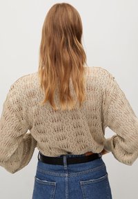 Femme aux cheveux longs châtain clair portant un pull beige à maille ajourée et un jean taille haute bleu avec une ceinture noire, debout de dos.