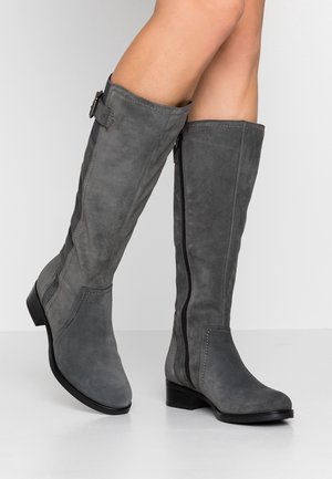 Bottes hautes en daim gris avec fermeture éclair latérale, petit talon et détail de boucle, portées sur des jambes croisées devant un fond uni.