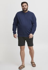Sudadera azul marino con cuello redondo y mangas largas, combinada con pantalones cortos negros y sandalias marrones, de pie sobre un fondo blanco.