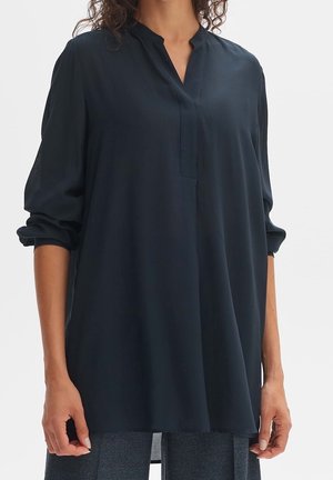 Femme portant une tunique ample bleu marine à manches longues avec un col mandarin et des manches retroussées, associée à un pantalon gris.