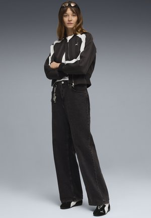 Schwarze, kurze Jacke mit weißen Streifen, kombiniert mit hochtaillierten schwarzen Jeans und schwarzem Schuhwerk. Auffälliges Kettendetail an den Jeans.