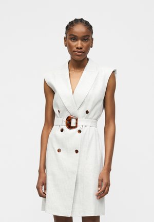 Žena nosiaca biele šaty bez rukávov s dvojradovým zapínaním, gombíky z korytnačky a zodpovedajúcim opaskom s veľkou prackou, stojaca pred jednotným pozadím.