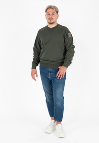 Felpa verde scuro con polsini e orlo a costine, caratterizzata da un collo rotondo e un piccolo logo applicato sulla spalla sinistra. Abbinata a jeans blu sbiaditi e sneakers bianche.