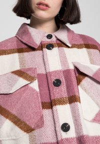 Rosa och krämfärgad rutig kappa med bruna detaljer. Gjord av ull, med krage, knäppningar och fickor med lock. Mjuk textur synlig.