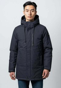 Homme portant un manteau d'hiver à capuche bleu marine avec fermeture éclair et boutons, se tenant contre un fond clair.