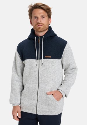 Veste à zip grise et marine avec une texture douce, une capuche, des poches avant et des blocs de couleur contrastants, mettant en vedette un accent en cuir brun.