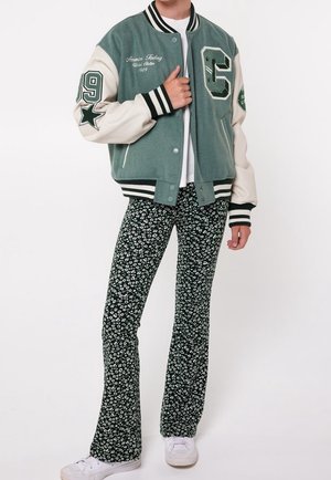 Giacca varsity verde e crema con patch ricamati e polsini a strisce, abbinata a pantaloni a campana floreali neri e sneakers bianche.