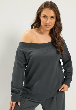 Kvinde med langt brunt hår iført mørkegrå off-shoulder sweatshirt og matchende bukser, poserer med den ene hånd hvilende på hoften.