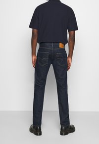 Man wearing dark blue jeans and black boots standing facing away against a plain light background.