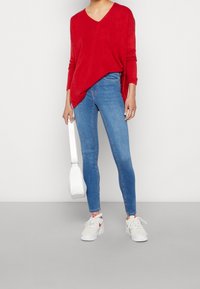 Czerwony sweter z dekoltem w kształcie litery V, niebieskie dopasowane dżinsy, biała torba na ramię i białe sneakersy z czerwonymi akcentami. Miękka tekstura, casualowy design.