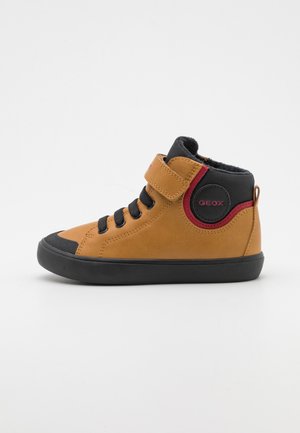 Zapato alto de cuero marrón con detalles en negro, cierre de cordones y velcro, suela de goma y un detalle de logo circular rojo en el costado.