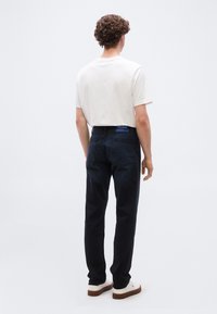 Νέος άνδρας με σγουρά μαλλιά, φορώντας λευκό μπλουζάκι, σκούρο μπλε τζιν με ετικέτα Hugo και λευκά αθλητικά παπούτσια, στέκεται γυρισμένος ανάποδα.