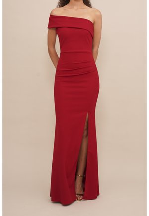 Vrouw draagt een vloerlang, off-shoulder rode jurk met een hoge split bij het dijbeen, staand met handen achter haar rug.
