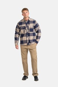 Camisa de franela con un gran patrón de cuadros en azul marino y beige, dos bolsillos en el pecho, cierre de botones en la parte delantera y mangas largas. Combinada con pantalones caqui y zapatos negros.