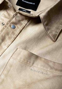 Chemise beige à boutons fabriquée en tissu texturé, dotée d'une fermeture à pression métallique, d'une poche cousue et d'un logo embossé sur la poche.