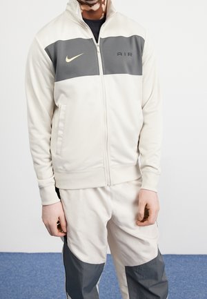Personne portant une veste zippée Nike Air crème et grise avec un pantalon assorti, debout contre un mur blanc et un sol bleu.