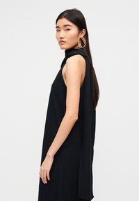 Μαύρο αμάνικο φόρεμα με ψηλό λαιμό, που διαθέτει ένα ρέον κάπα λεπτομέρεια και λεία υφή. Χοντρά χρυσά κρίκοι σκουλαρίκια ολοκληρώνουν την εμφάνιση.