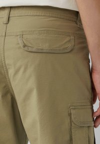 Khaki cargo-housut, joissa on teksturoitu pinta, sisältävät takataskun pyöristettyine reunoineen ja vahvistetut saumat. Sivutasku näkyvissä.