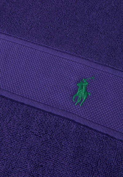 Κοντινό πλάνο σε μοβ υφασμάτινη υφή με μια οριζόντια λωρίδα που φέρει ένα μικρό πράσινο κεντημένο λογότυπο παίκτη πόλο.