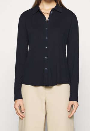Vrouw draagt een zwart lang mouw knoopblouse en beige broek, met een gouden ketting zichtbaar rond haar hals.