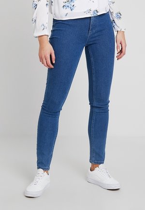 Jeans de denim azul ajustados con una textura suave, corte de altura media y un diseño minimalista, combinados con zapatillas blancas.