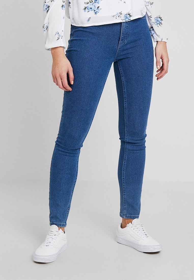 Jeans de denim azul ajustados con una textura suave, corte de altura media y un diseño minimalista, combinados con zapatillas blancas.