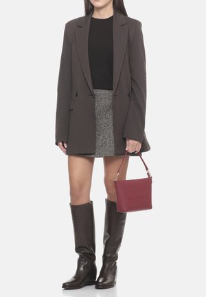 Donna con blazer marrone scuro, top nero, gonna grigia, stivali marrone scuro fino al ginocchio, che tiene una piccola borsa rossa per la cinghia.