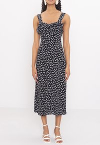 Černé šaty s bílým květinovým vzorem, volánkovým výstřihem a širokými ramínky. Kolenové délky s bočním rozparkem. Oblečené s bílými sandály na podpatku.