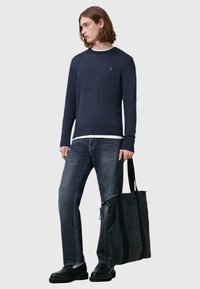Marineblauer Pullover mit Rundhalsausschnitt, dunkle Jeans und schwarze Ledertasche. Das Model hat lange Haare und trägt schwarze Stiefel. Schlichtes, minimalistisches Design.