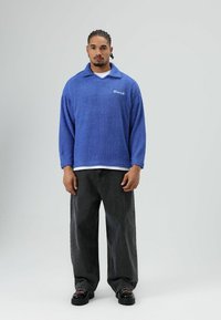 Blauwe fleece pullover met kraag en logo, gecombineerd met losse zwarte jeans en stevige zwarte schoenen. Gestructureerde, zachte stofafwerking.