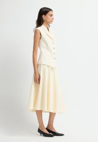 Kremfarvet, ærmeløs, skræddersyet vest med gyldne knapper, kombineret med et flydende, knælange skørt i samme nuance og sorte spidse flade sko.